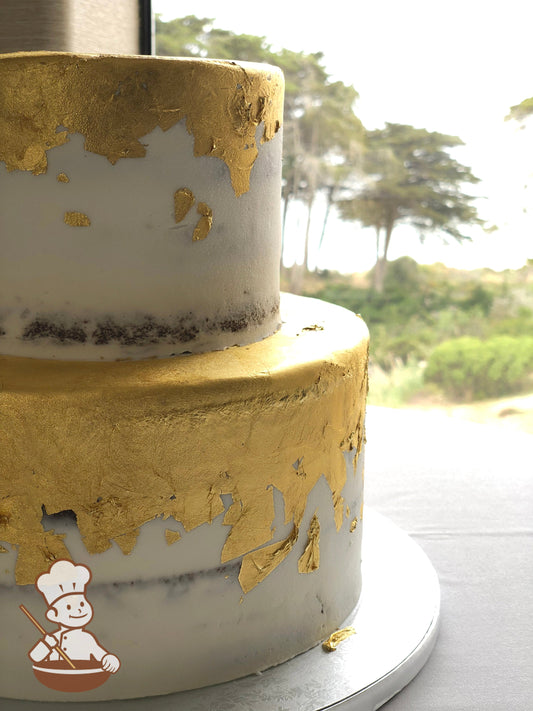 2-tier cake with white icing & decorated with a scraped texture that shows some of the cake and gold sheets on the top of the tiers coming down.