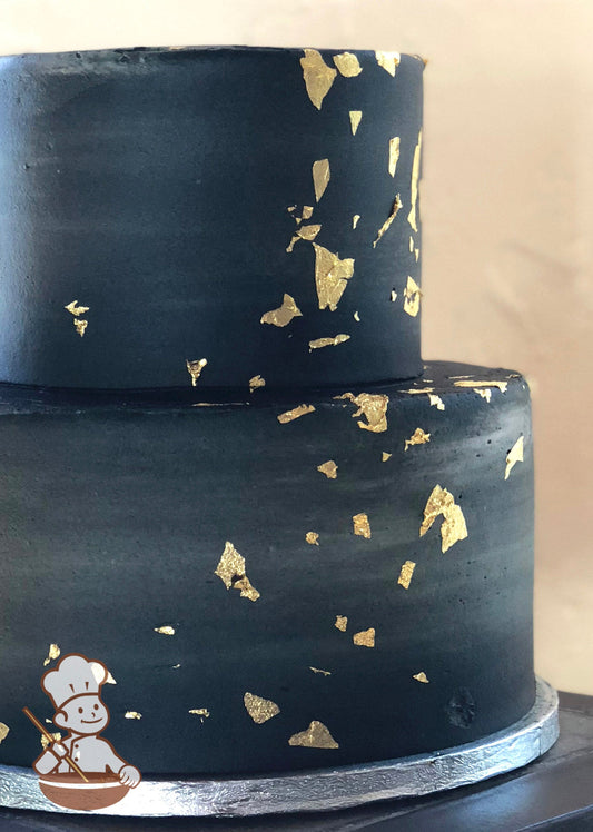 2-tier cake with black smooth icing and decorated with gold foil flakes.