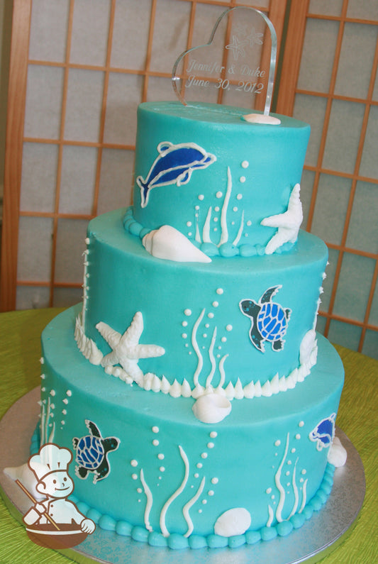 3-tier blue cake with piped turtles, dolphins, kelp and ocean bubbles. Decorated with seashells.
