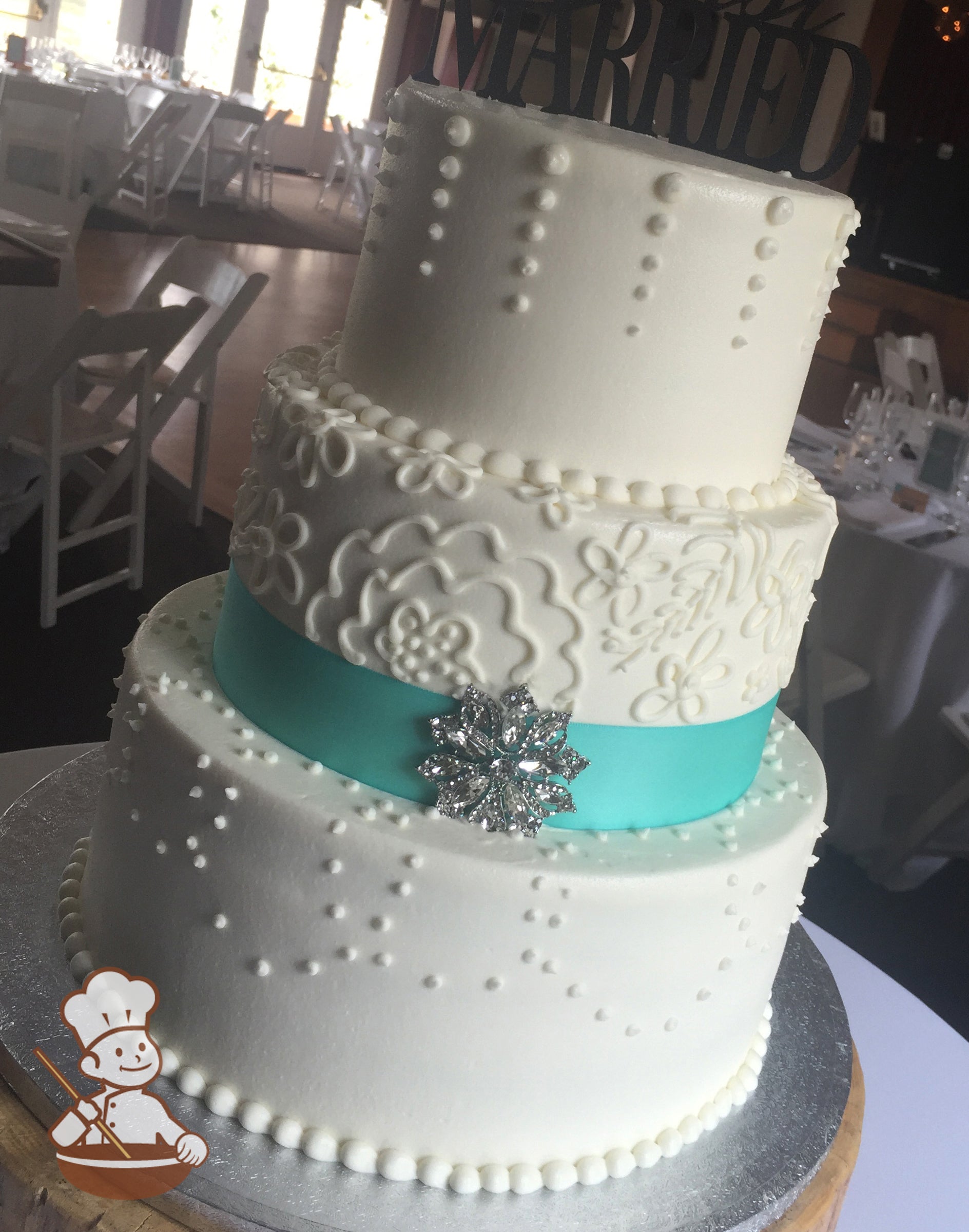 3-tier cake with smooth white icing and decorated with different white buttercream piping's and a turquoise ribbon with a silver brooch.