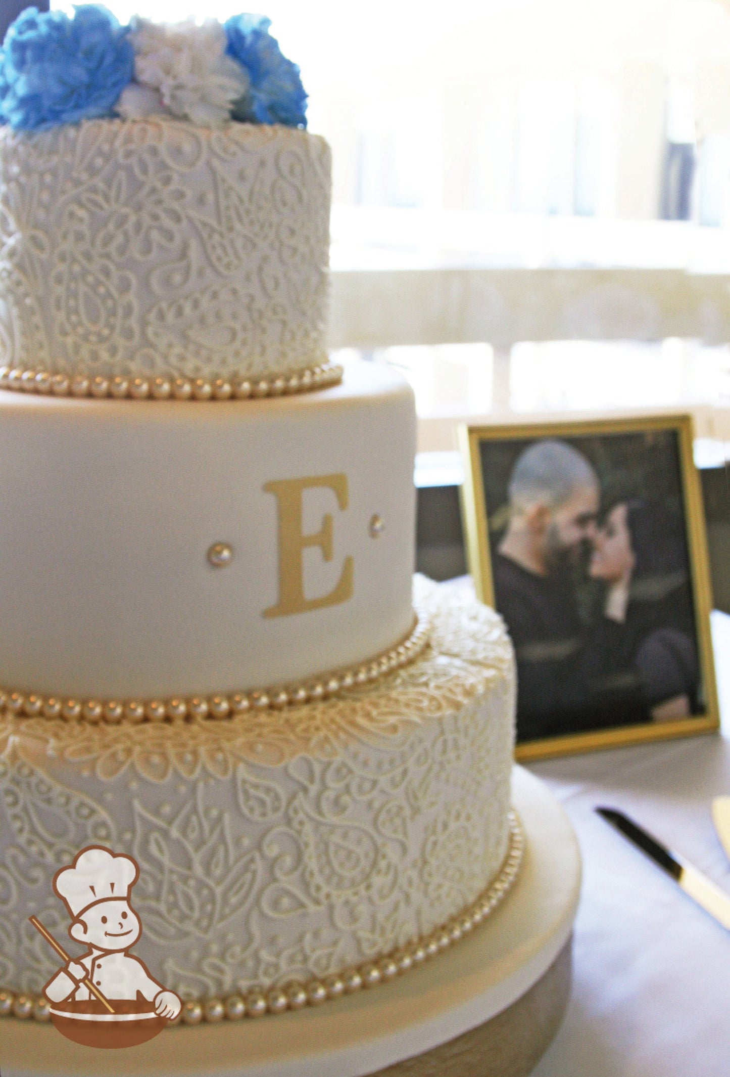 3-tier cake with smooth white icing and decorated with buttercream piped flowers and scrolls on bottom and top tier and a fondant monogram.