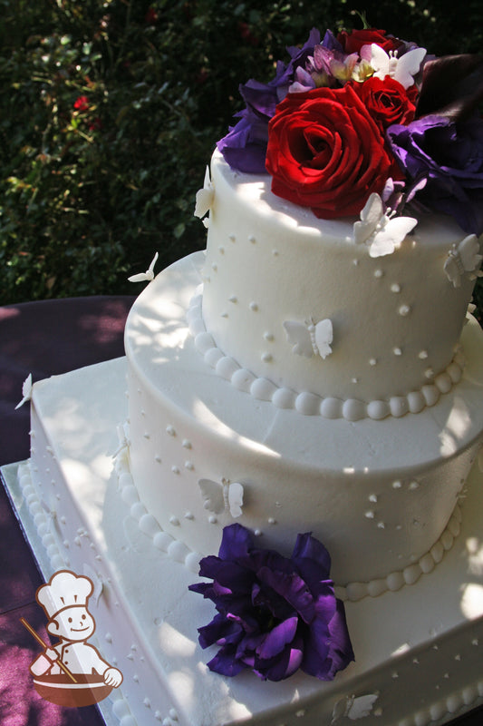 3 tier wedding cake with square bottom tier and round top tiers decorated with sugar butterflies and fresh flowers in reds, purples and pinks.