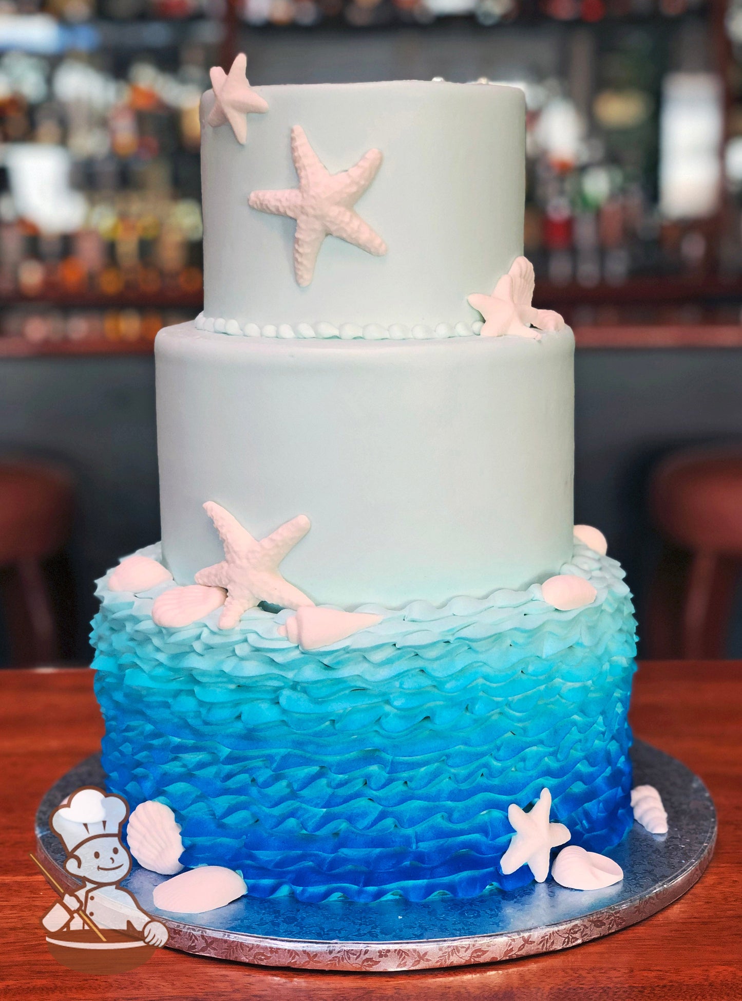 Cake decorated with wavy buttercream piping on the bottom tier, smooth light-blue icing on the middle and top tier and white fondant shells.