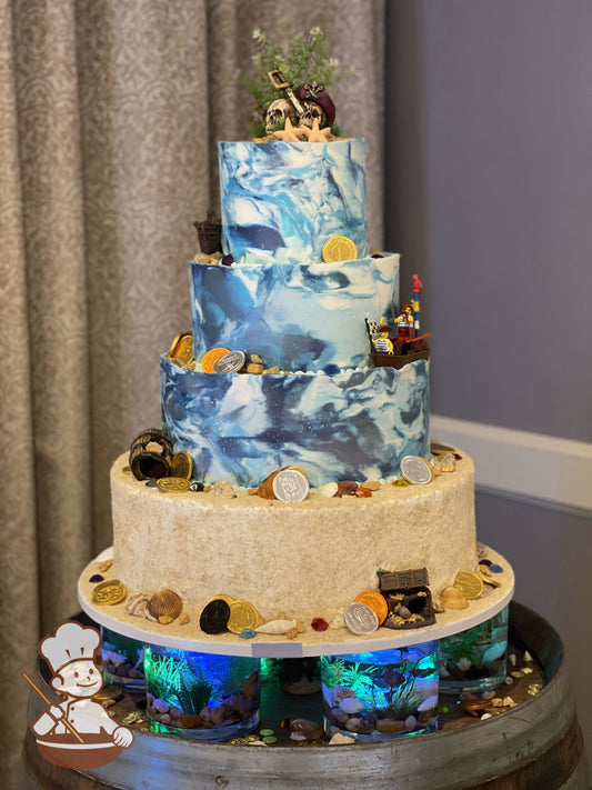 4-tier cake with bottom tier covered in sanding sugar and top three tiers in blue marble buttercream. Decorated with seashells and pirate toys.