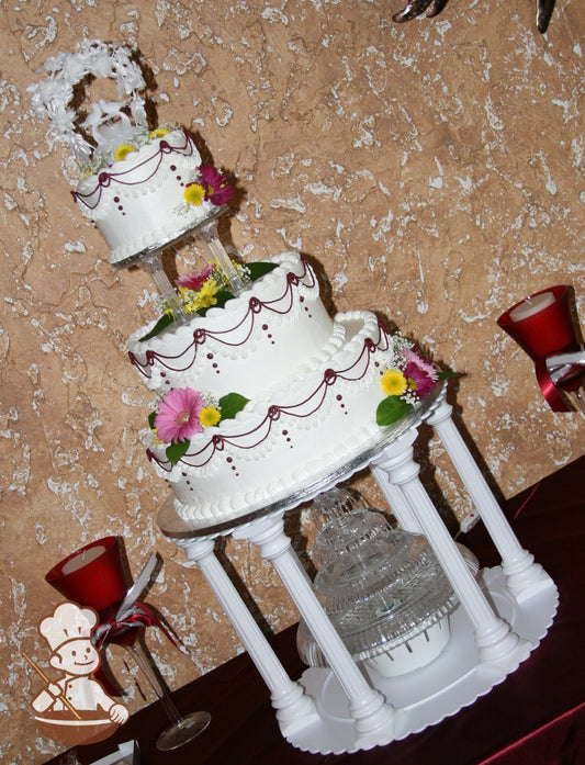 3-tier white cake with white and burgundy drapes piped around all tiers. Plastic pillars surround a water fountain and holding up the cake.