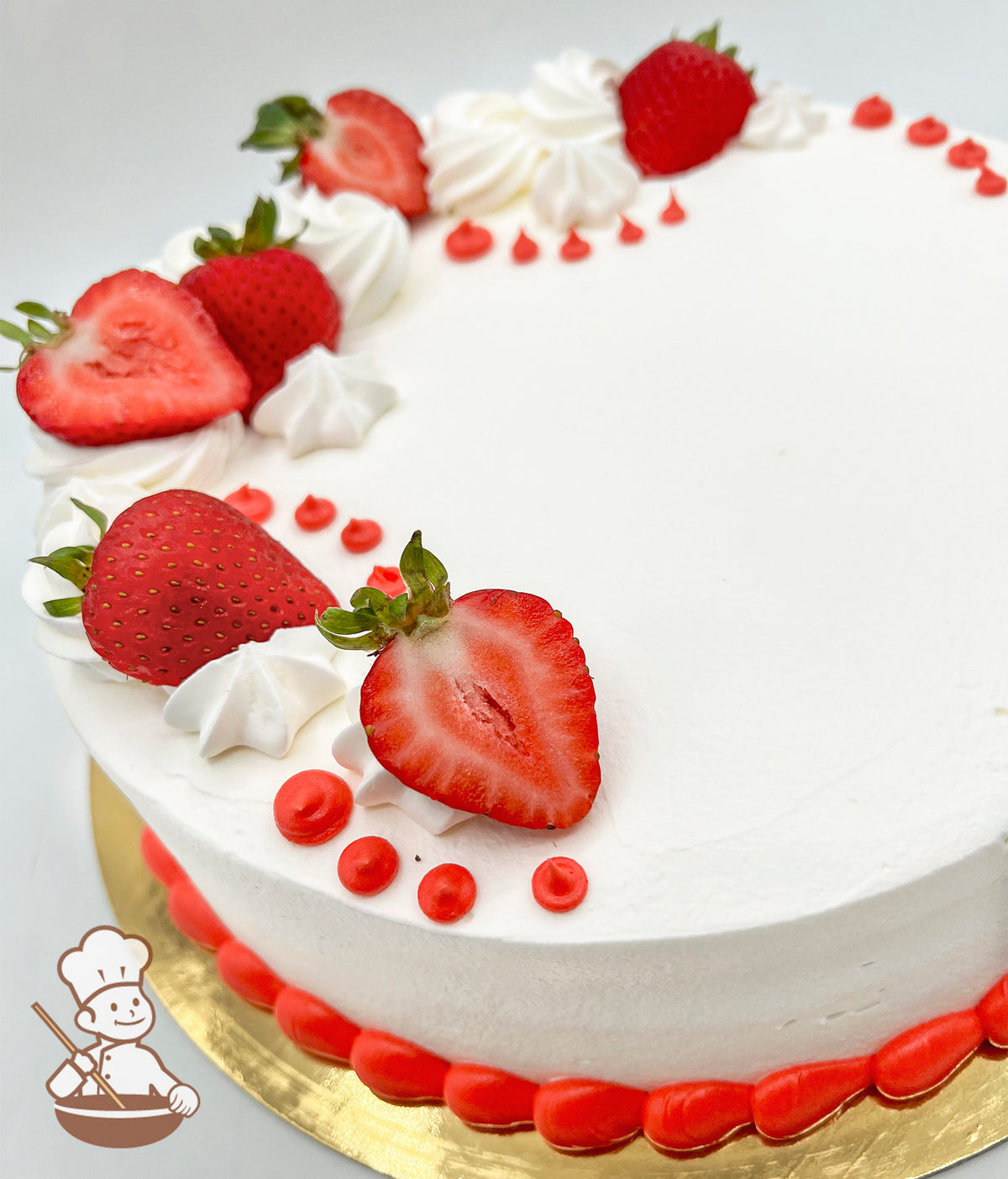White frosted cake topped with fresh strawberries, dollops of white buttercream, and small red icing dots, on a gold cake board.