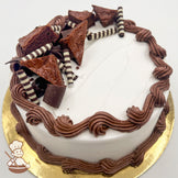 Round cake with white frosting, brown buttercream borders, topped with chocolate brownie triangles, striped chocolate sticks, and thick brown chocolate curls.