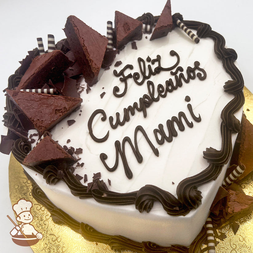 Heart-shaped cake with white frosting, brown buttercream borders, triangular chocolate brownie pieces, striped chocolate sticks, and thick brown chocolate curls on a gold base.