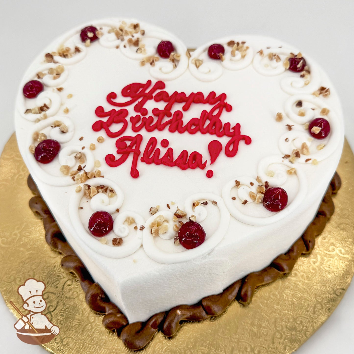 Heart-shaped white iced cake with red raspberry jam accents, swirled frosting details, and sprinkled nuts on top, placed on a gold cake board.