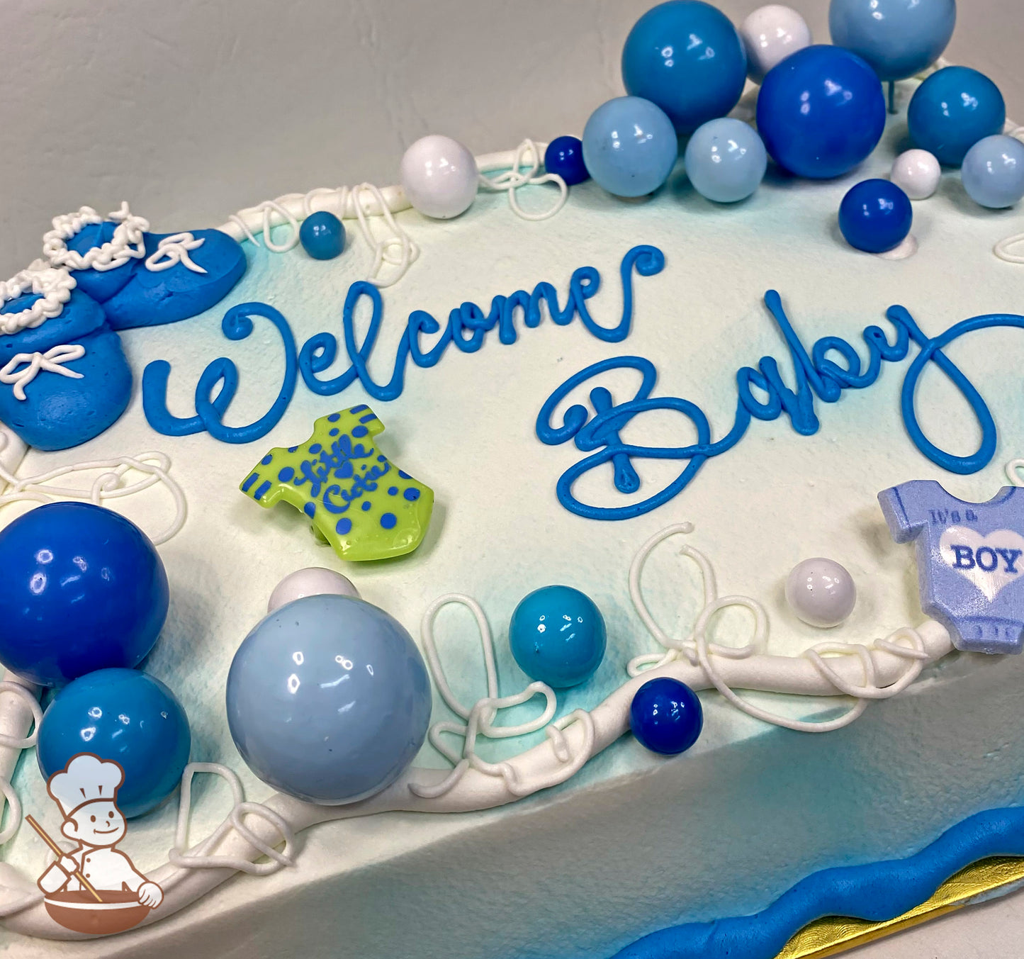 Sheet cake with blue baby booties, bubble toppers, and baby rings. Decorated with blue and white buttercream trims and plastic baby-themed toys.