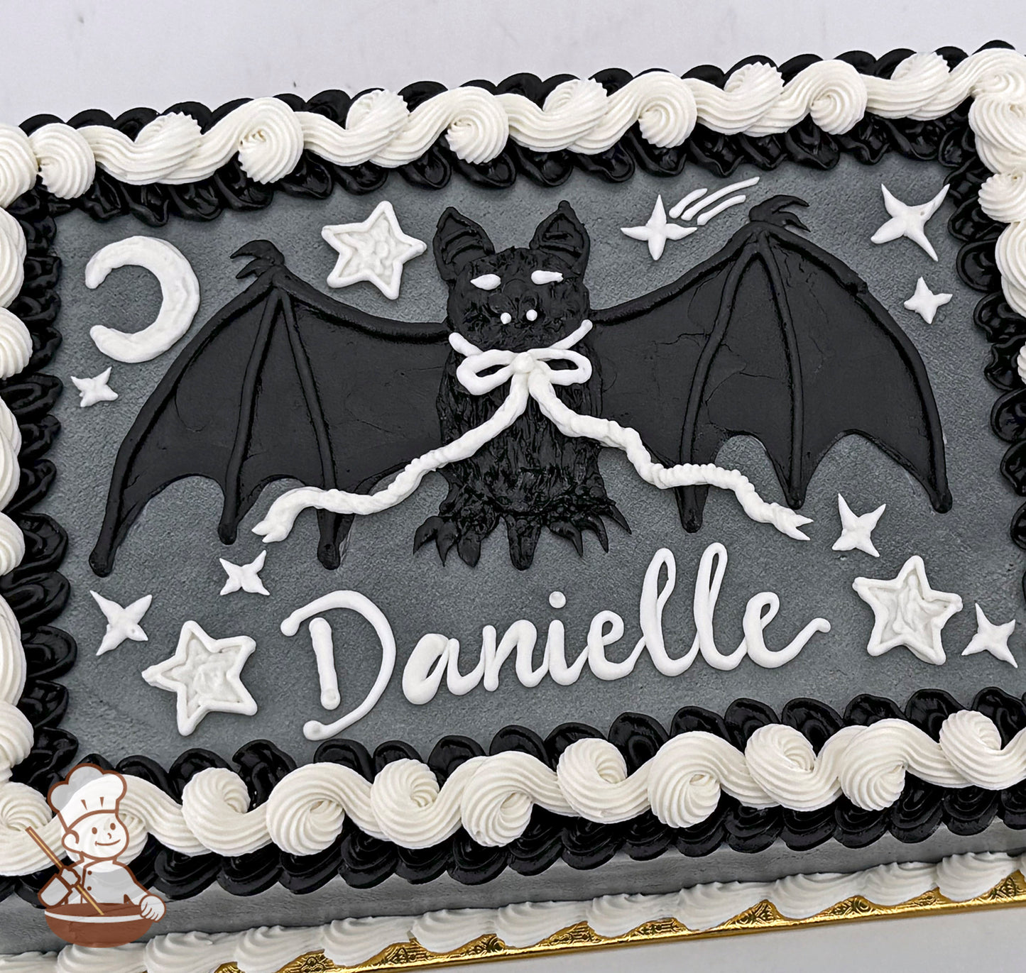 A sheet cake with a black bat in the center, surrounded by white buttercream stars and a crescent moon. The border is swirled black and white.