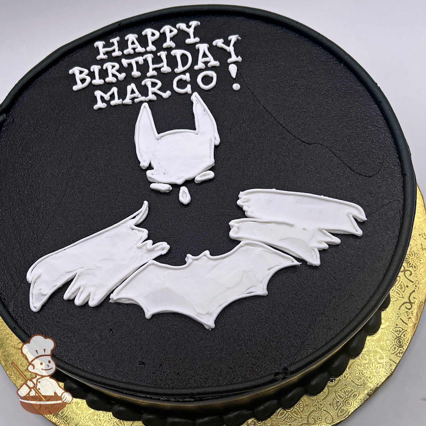 Round cake with black base, white buttercream Batman outline featuring bat wings and mask, gold decorative cake board.