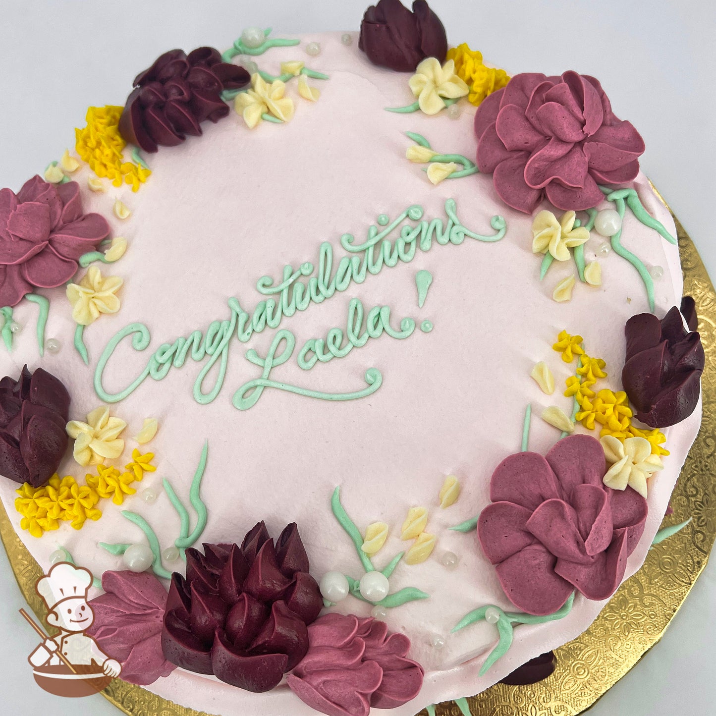 Round cake with pink background, decorated with burgundy bell flowers, pale cream blossoms, yellow florals, green stems, and white sugar pearls.