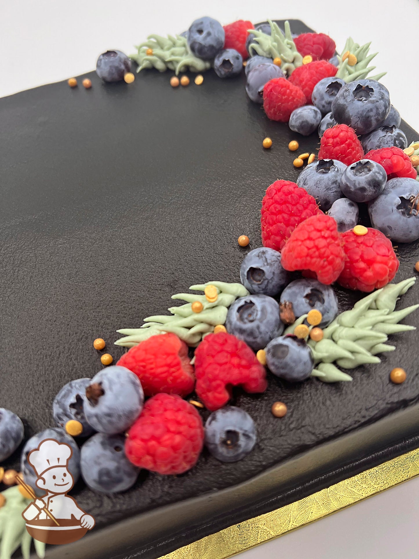 Sheet cake with black icing, decorated with fresh blueberries, raspberries, green buttercream leaves, and gold sugar pearls.