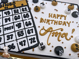 Sheet cake with buttercream bingo card and balls, decorated in black, white, and gold. Gold, black and white buttercream dots scattered around.