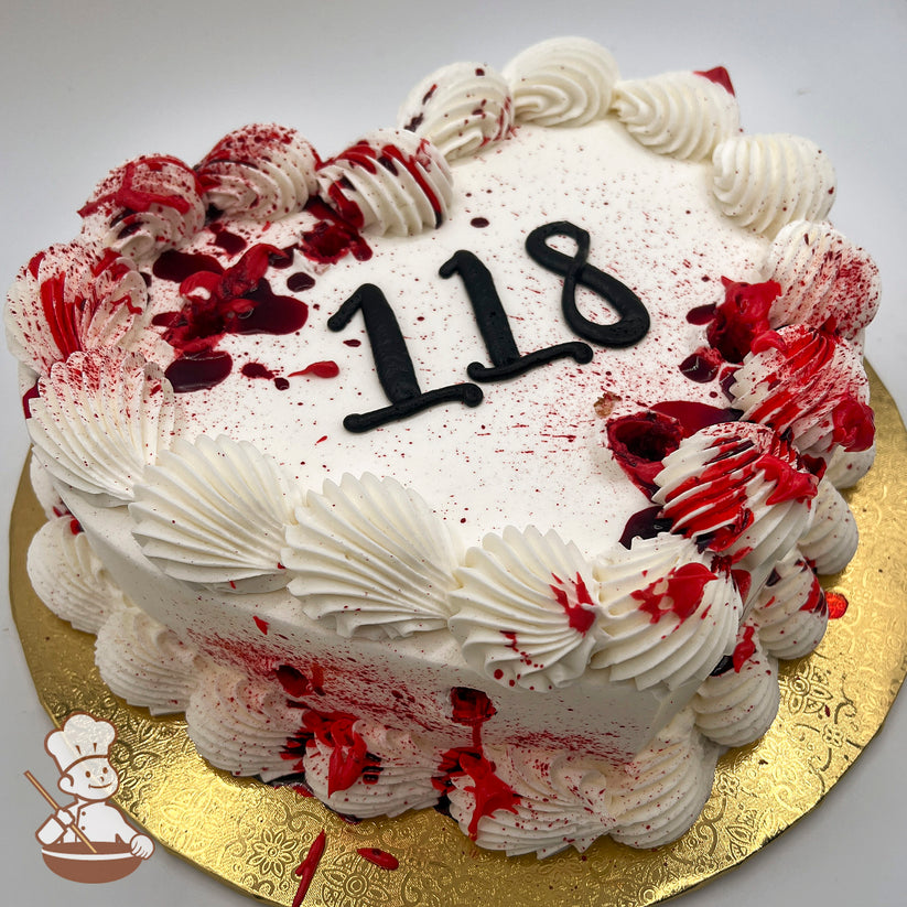 White heart cake with piped buttercream swirl trims along the edges, splattered with red coloring resembling blood stains, on a gold board.