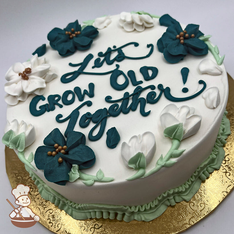 Round white cake with buttercream decorations featuring dark teal blossoms, white bell flowers, green leaves, and a ruffled green border.