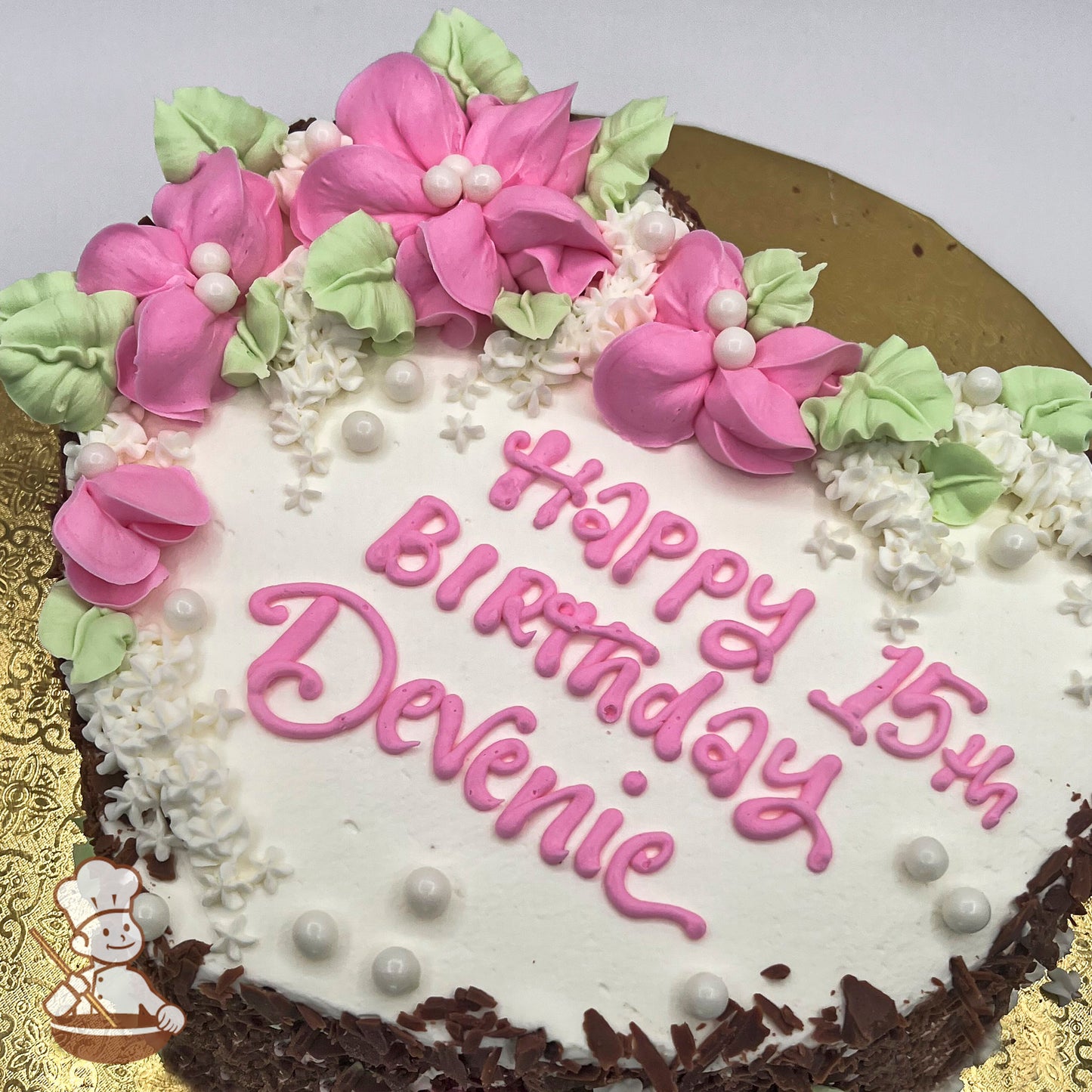 Round cake with pink buttercream blossoms, green leaves, white sugar pearls, and small white flowers on a white frosting base.