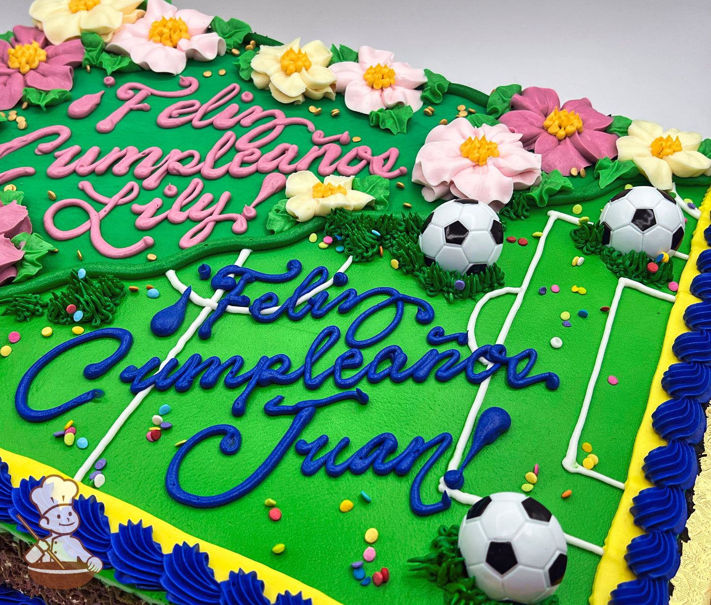 Sheet cake with green background resembling a soccer field, decorated with plastic soccer ball toys, pink and yellow buttercream flowers, and blue border piping.