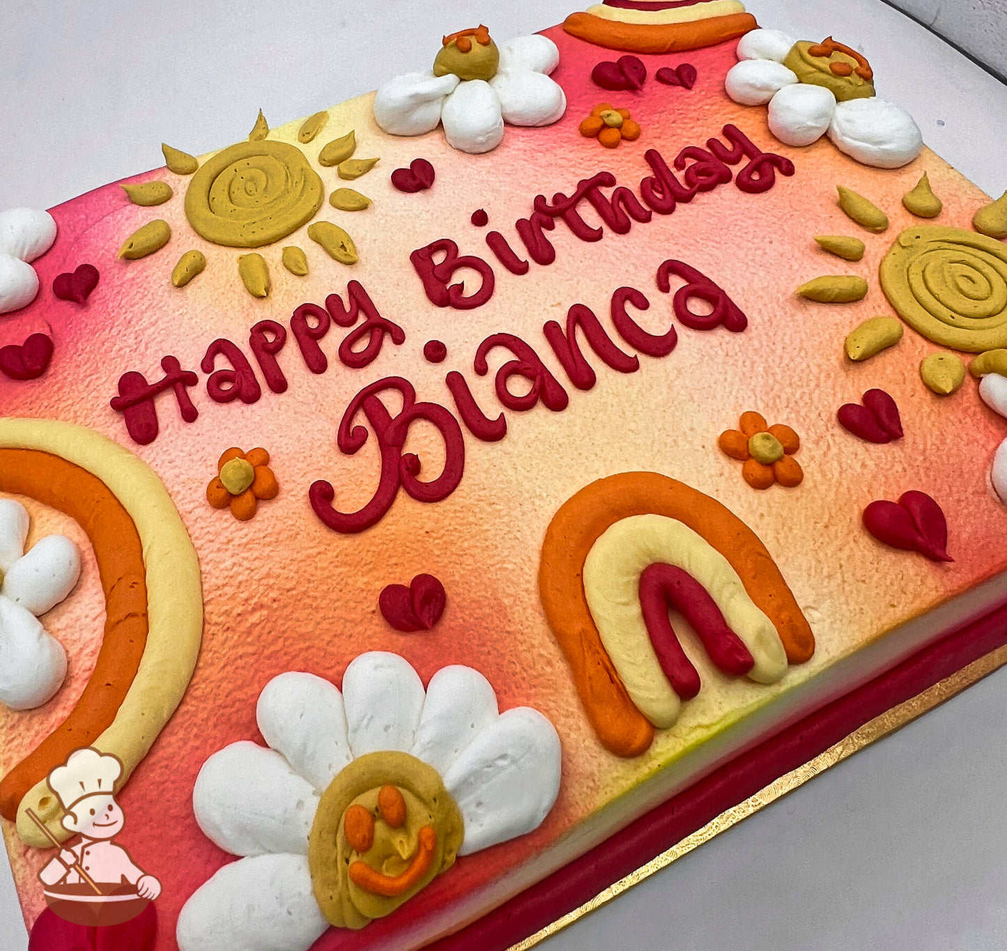 Sheet cake with ombre pink and orange background, decorated with rainbows, smiling daisies, yellow sun, small orange flowers, and red hearts.