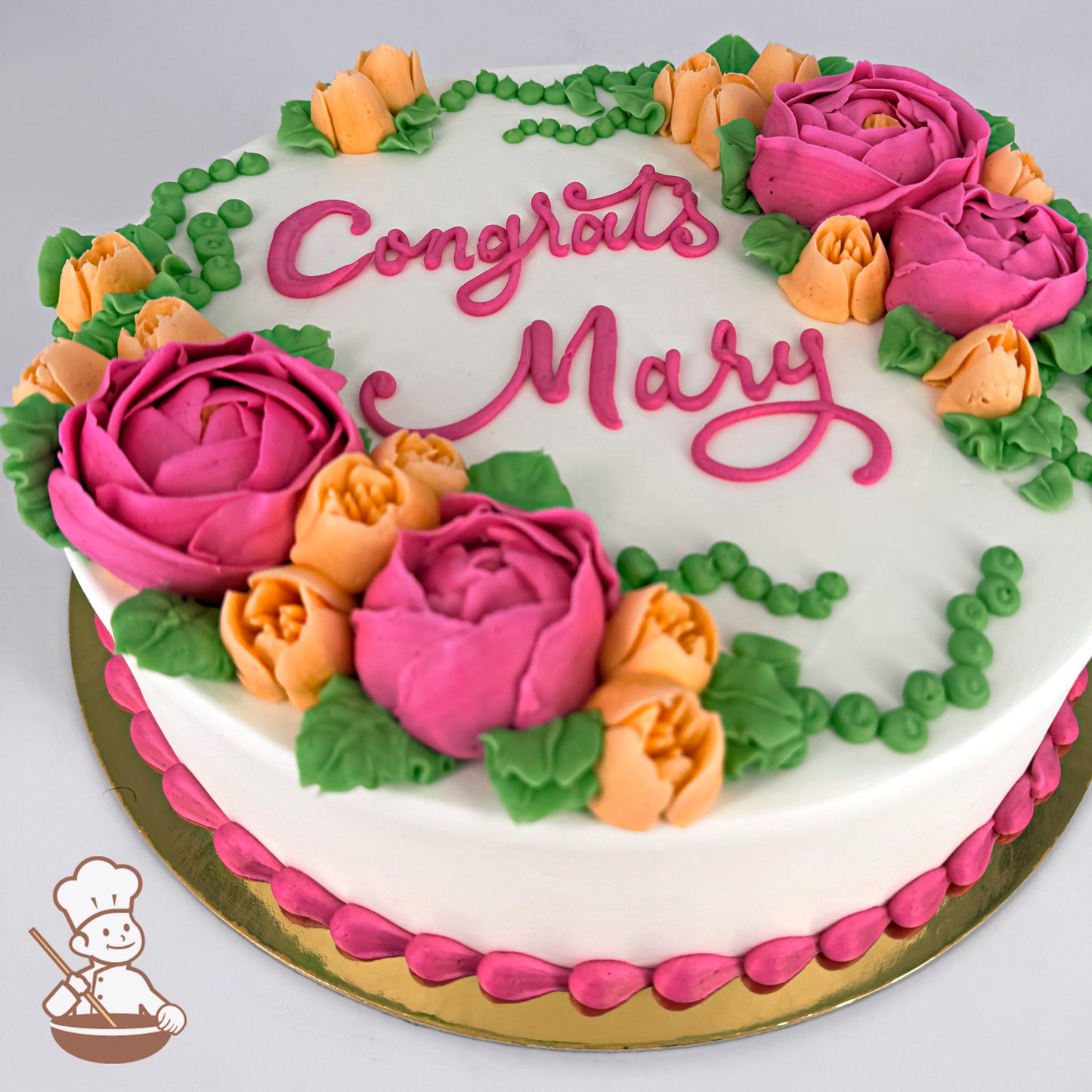 Round cake with buttercream decorations featuring pink buttercups, orange freesia, green string of pearls, and leaves. Pink border accents the base.
