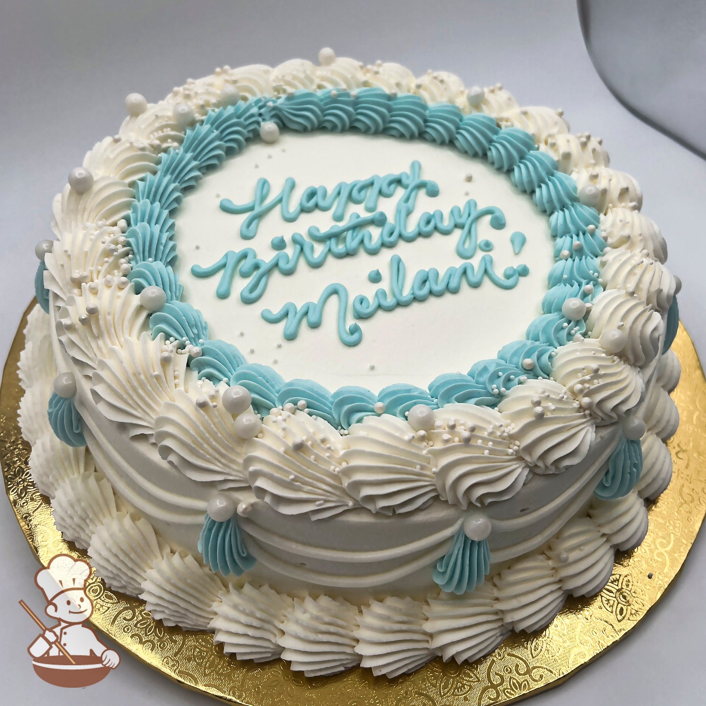 Round white cake with blue and white piped swirls, sugar pearls, and vintage-style trims on a gold board.