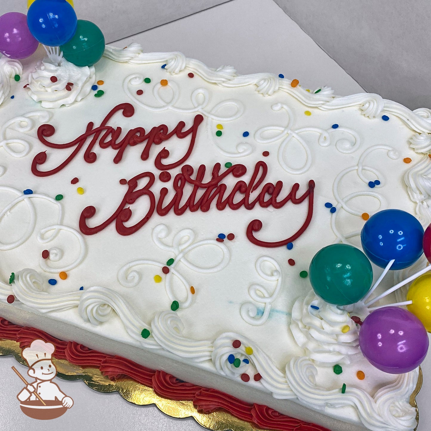 Sheet cake with white frosting, colorful confetti sprinkles, decorative scroll piping, and plastic balloon toppers in blue, green, purple, and yellow.