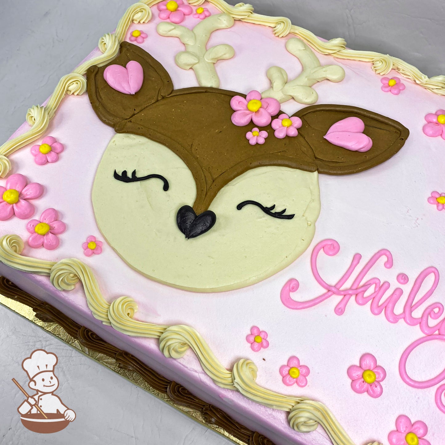 Sheet cake with buttercream decorations featuring a cute deer face, antlers, pink heart accents, and pink and yellow daisies on a pastel background.