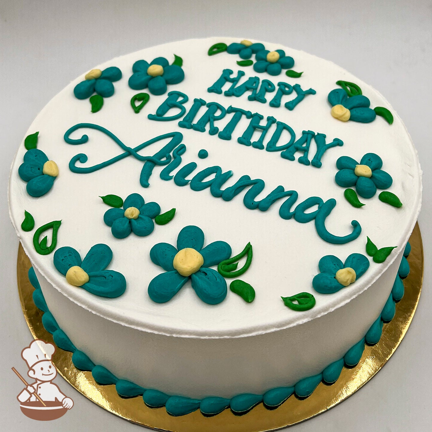 Round white iced cake decorated with teal daisy flowers, yellow centers, and green leaves. Teal piped border on a gold cake board.
