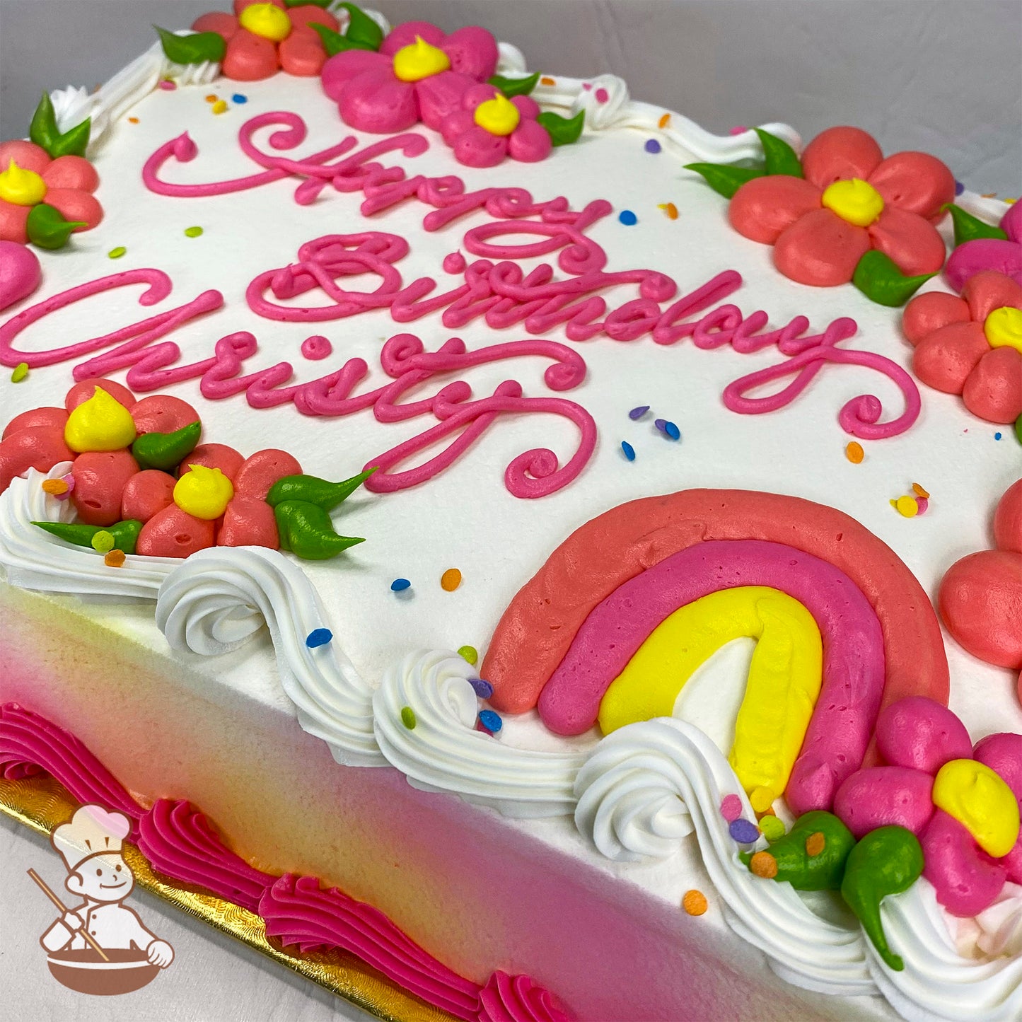 Sheet cake with white frosting, decorated with pink and yellow daisies, a pink and yellow buttercream rainbow, colorful sprinkles, and white swirl trims.
