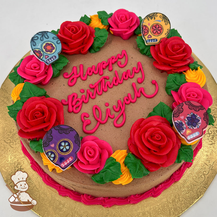 Round cake with brown icing, decorated with hand-piped red, pink, and yellow roses, green leaves, and colorful plastic sugar skull rings on a gold base.