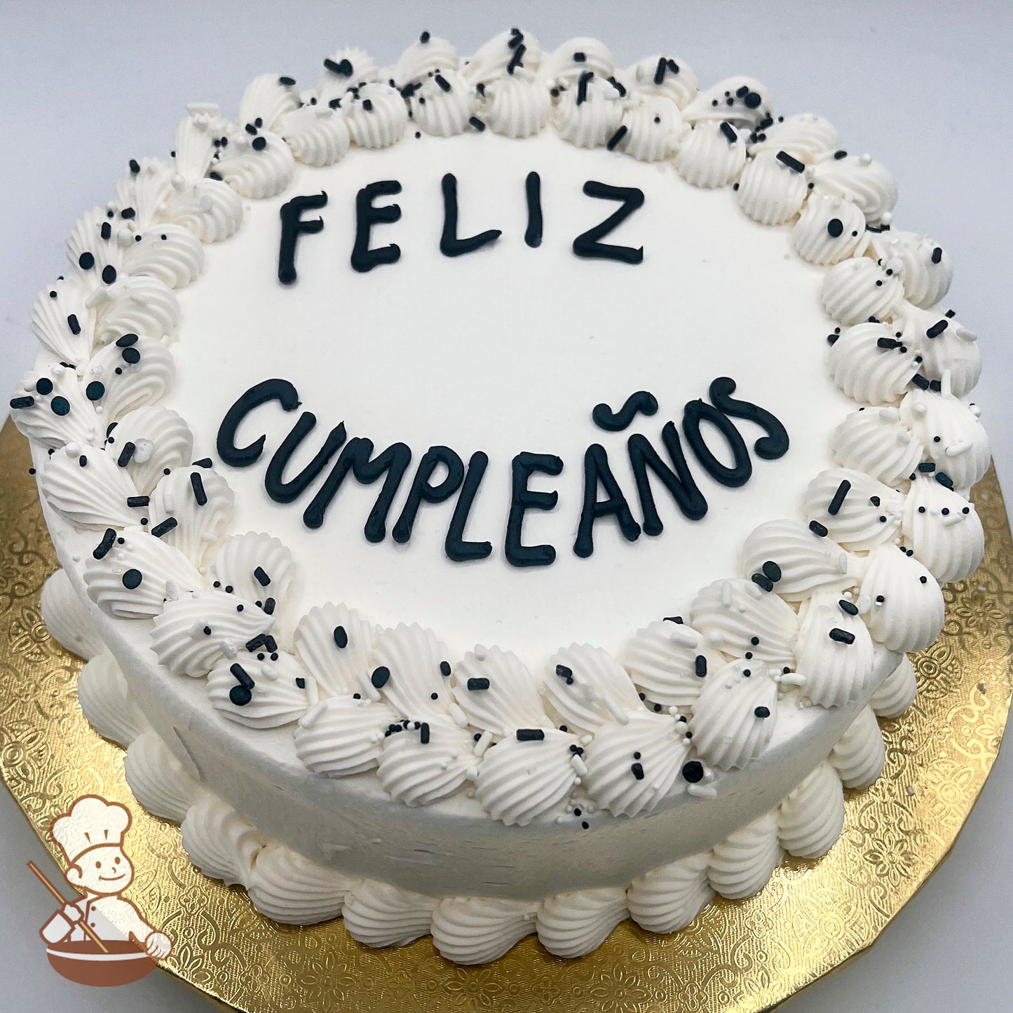 Round cake with white icing, decorated with hand-piped double scallop trim and black sprinkles on top, on a gold board.