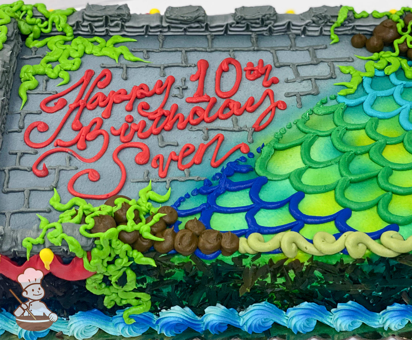 Sheet cake with a gray buttercream castle wall design, green vines, colorful hand-piped dragon scales in blue and green and blue waves at the base.