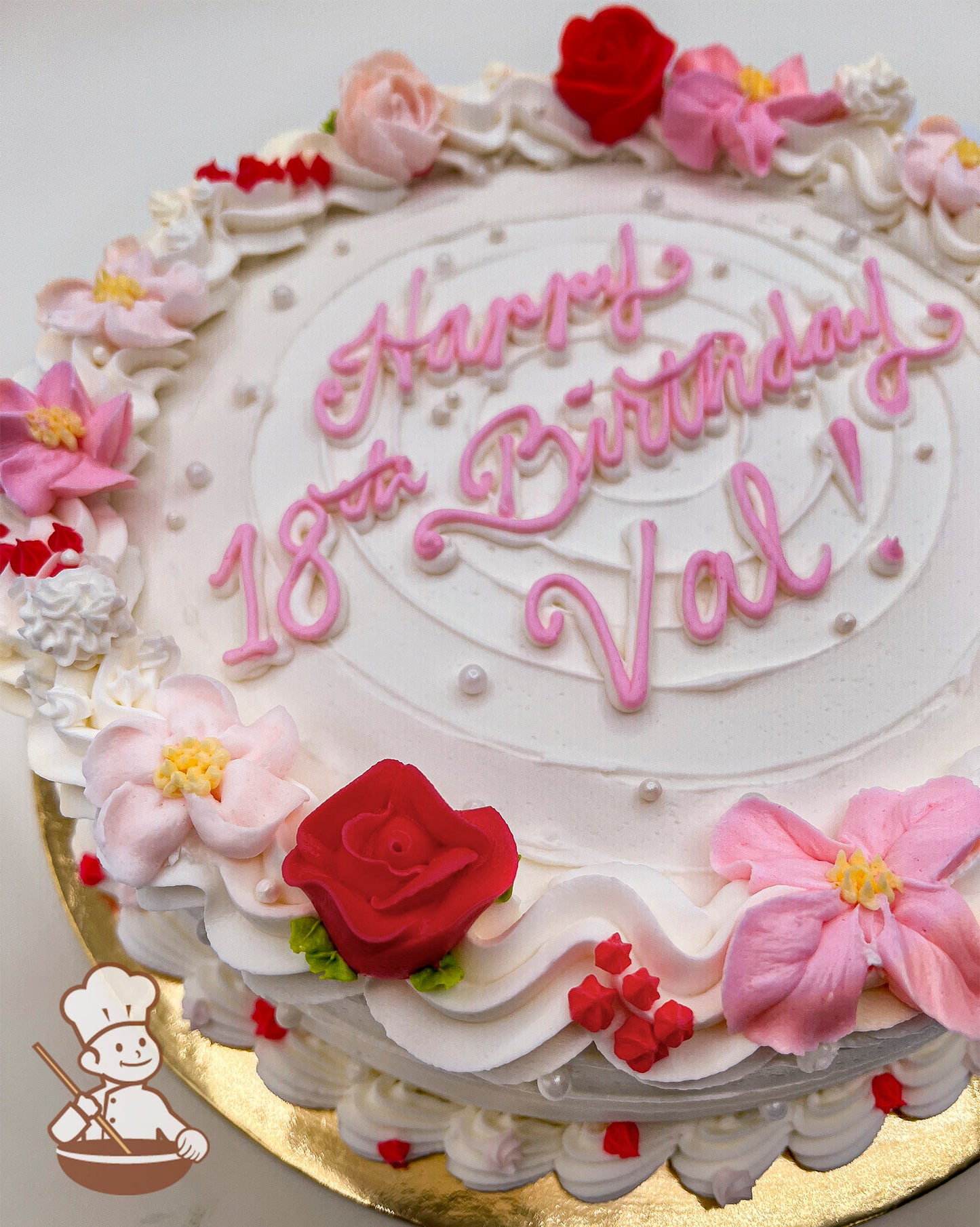 Round cake with white frosting, decorated with pink and red buttercream flowers, small pearls, and piped swirl trims.