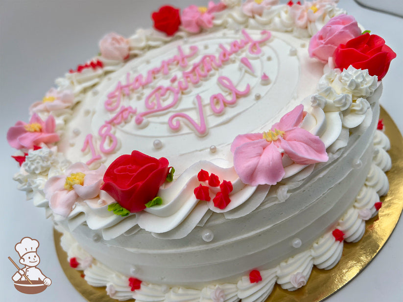 Round cake with white frosting, decorated with pink and red buttercream flowers, small pearls, and piped swirl trims.