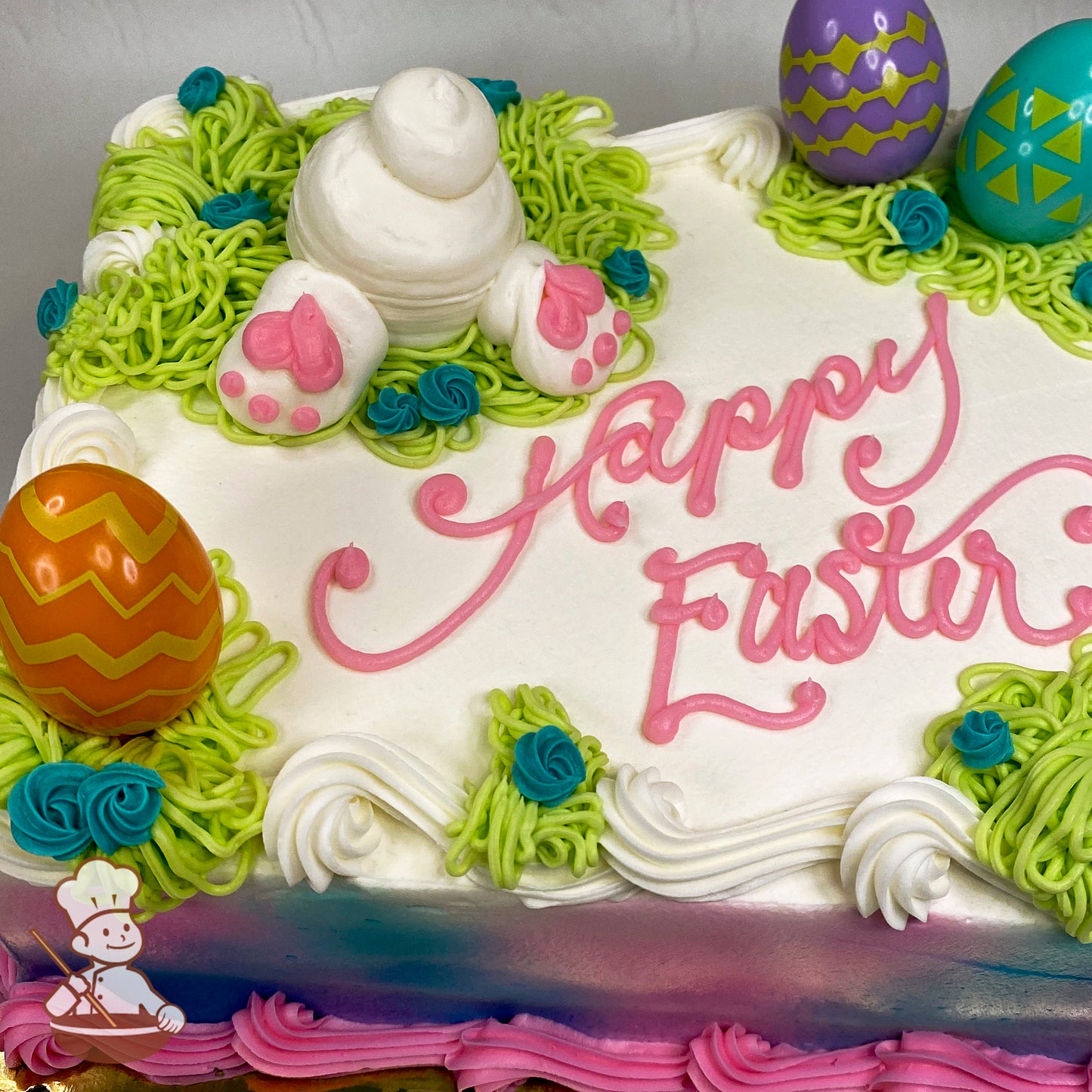 Sheet cake with buttercream bunny tail, surrounded by green grass, plastic Easter egg rings in orange, purple, and teal, and blue floral accents.