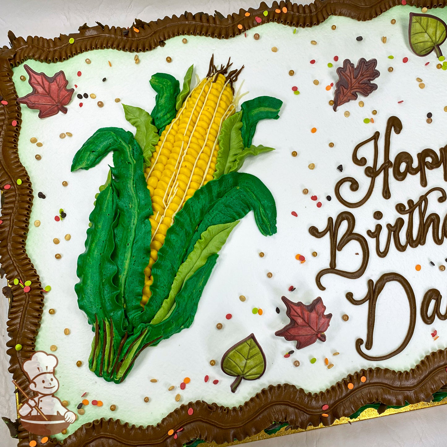 Sheet cake with a hand-piped buttercream corn design, green leaves, brown borders, plastic leaf decorations, and scattered fall-colored sprinkles.
