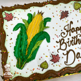 Sheet cake with a hand-piped buttercream corn design, green leaves, brown borders, plastic leaf decorations, and scattered fall-colored sprinkles.