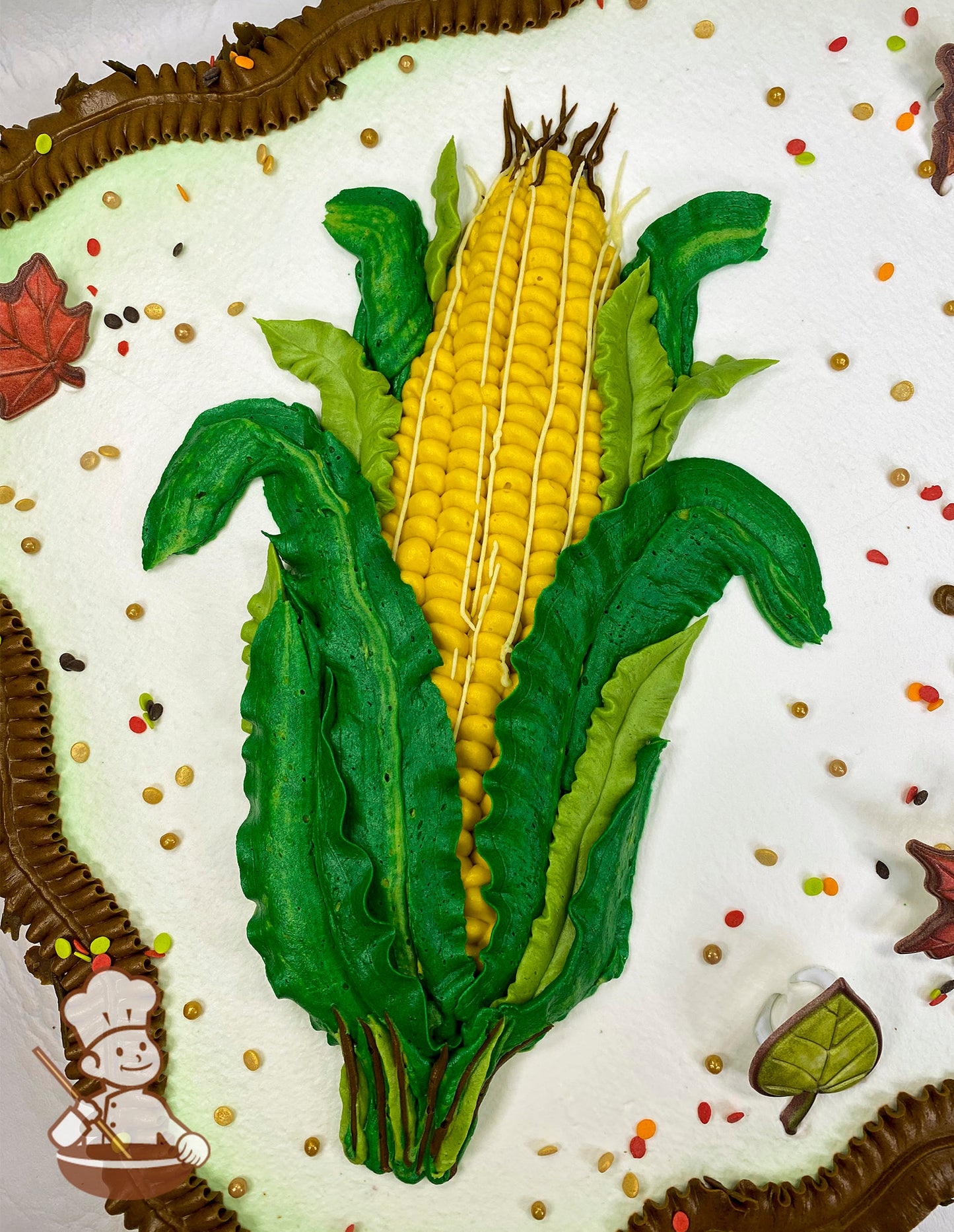 Sheet cake with a hand-piped buttercream corn design, green leaves, brown borders, plastic leaf decorations, and scattered fall-colored sprinkles.