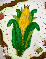 Sheet cake with a hand-piped buttercream corn design, green leaves, brown borders, plastic leaf decorations, and scattered fall-colored sprinkles.