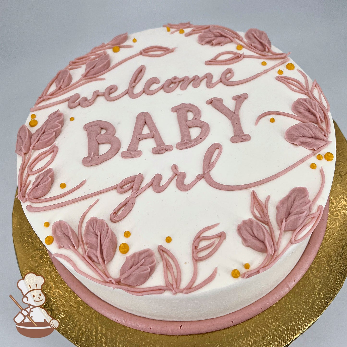 Round cake with white frosting, decorated with pink hand-piped ferns, leaves, and small yellow dots. Placed on a gold embossed board.
