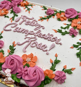 Sheet cake with white frosting, decorated with hand-piped pink rosettes, orange flowers, green vines, and leaves.