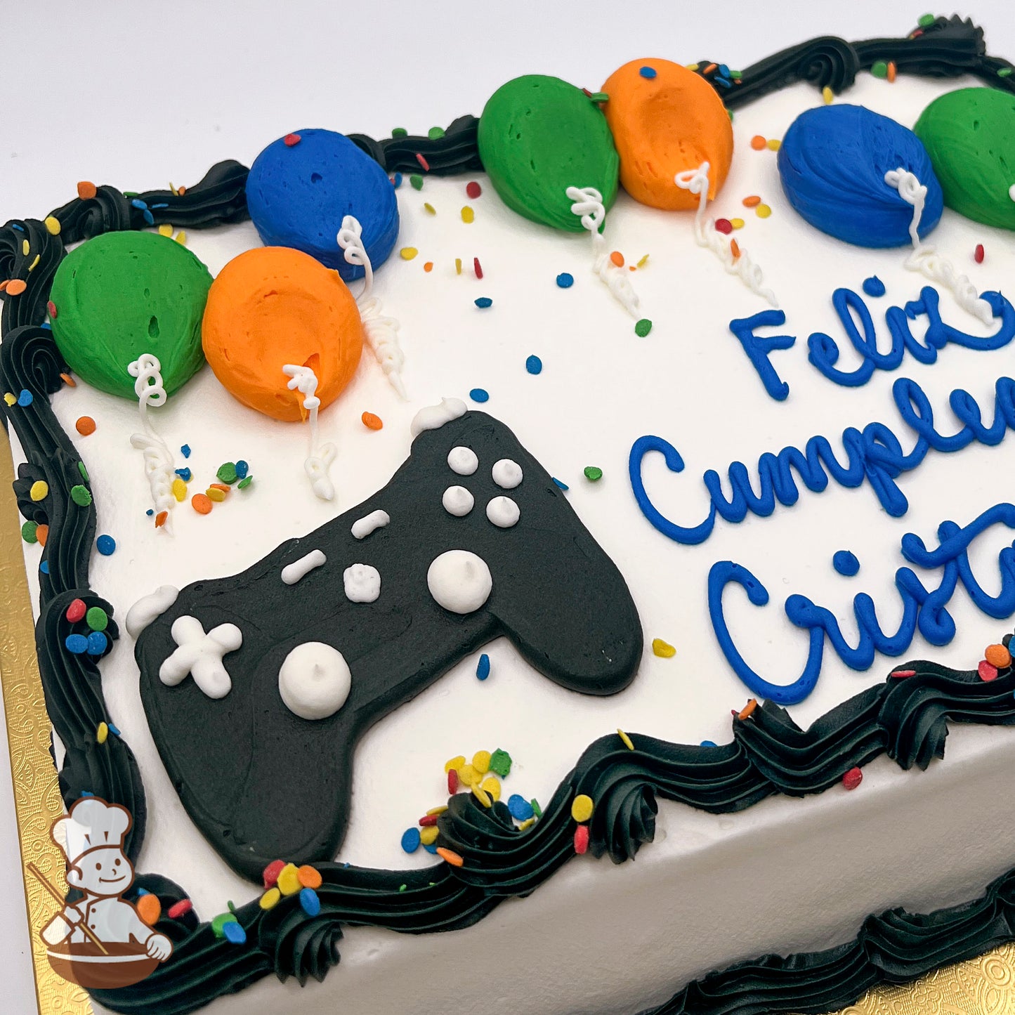 Sheet cake with icing decorations, featuring a black game controller design and colorful balloons in green, orange, and blue, with confetti accents.