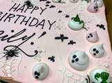 Pink sheet cake with buttercream ghosts, black bats, white spider webs, plastic spiders, and scattered white sugar pearls for decoration.