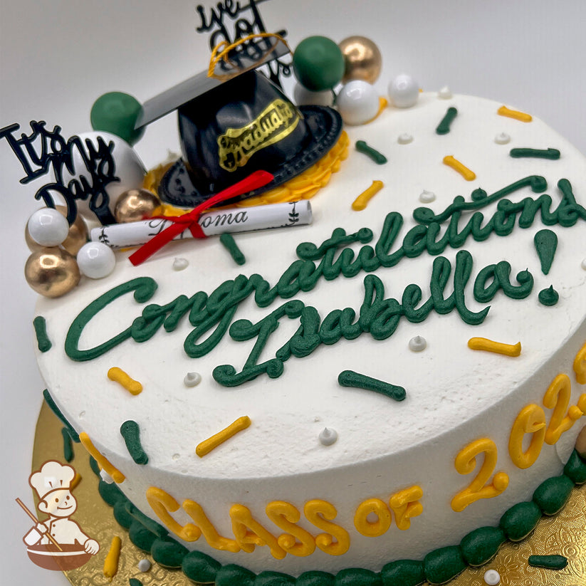 Round cake with white frosting, green and yellow buttercream sprinkles, a plastic black grad cap, a paper diploma with a red ribbon, and gold, green, and white bubble toppers.