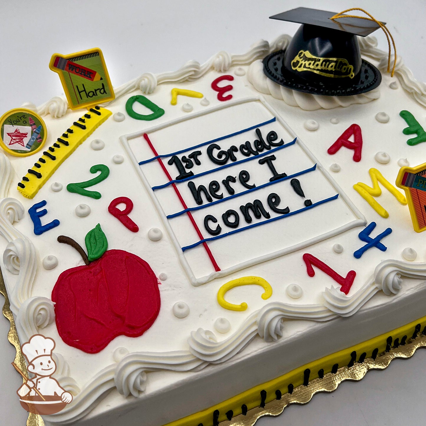 Sheet cake with white frosting, decorated with a plastic graduation cap, red apple, ruler, colorful letters, numbers, and dots.