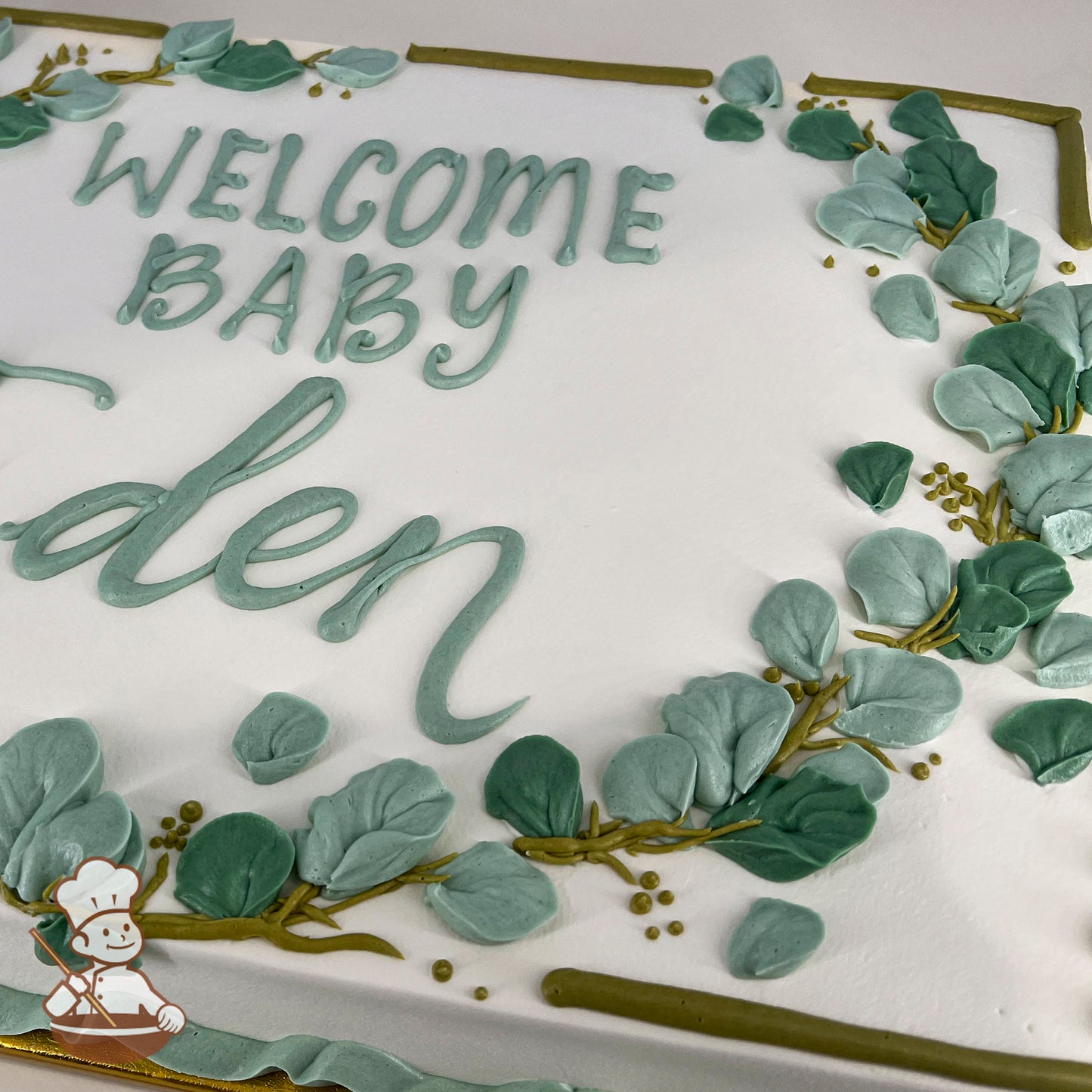 Sheet cake with white frosting, decorated with green buttercream eucalyptus leaves, golden branches, and a piped message in green icing.