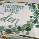 Sheet cake with white frosting, decorated with green buttercream eucalyptus leaves, golden branches, and a piped message in green icing.