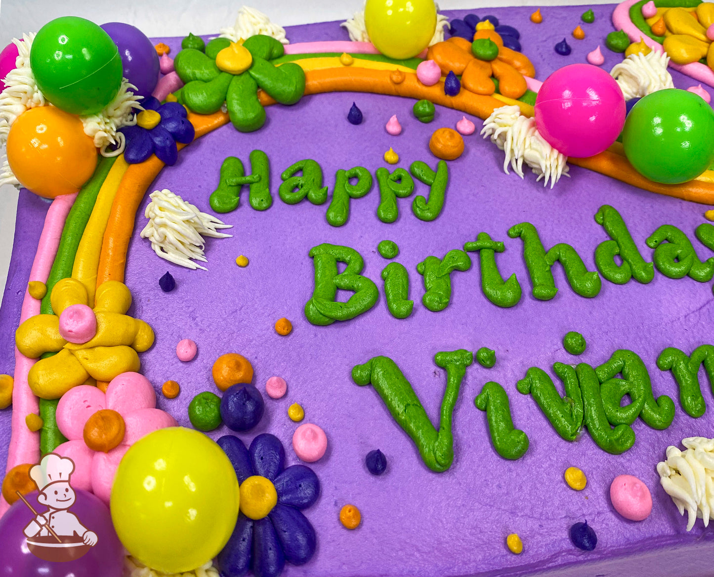 Purple sheet cake with colorful buttercream decorations, including daisies, a rainbow, dots, and plastic balloons in green, yellow, pink, and orange.