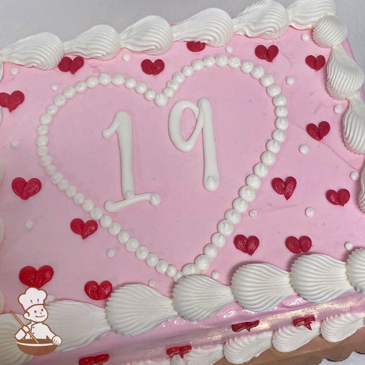 Pink sheet cake with a white buttercream heart frame outlined by dots, red heart decorations, and scalloped white buttercream trim along the edges.